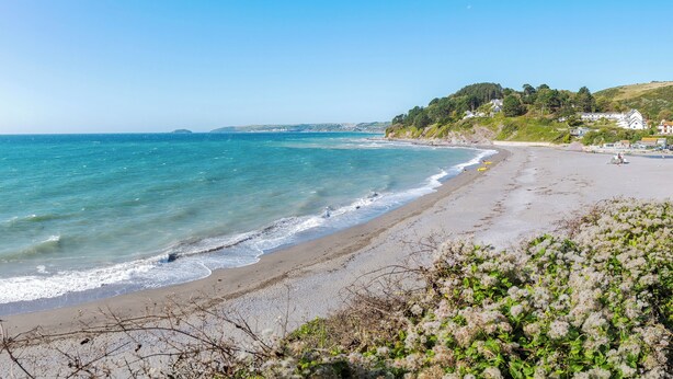 En strand med kristallklart blått vatten, en sandstrand och hus som ligger på en sluttning i bakgrunden.