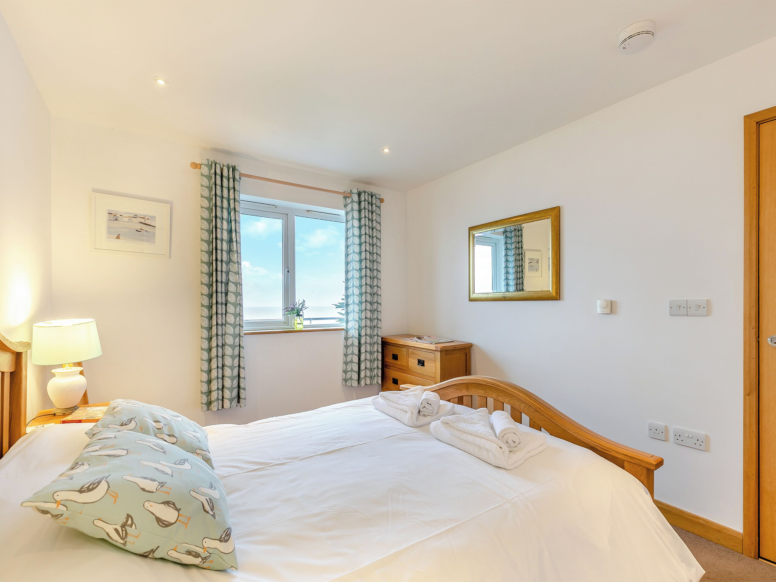 A bedroom with a bed, a bedside table with a lamp, a window with curtains, a mirror, and a door.