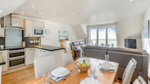 Uma cozinha moderna com eletrodomésticos embutidos, uma sala de jantar com mesa de madeira posta para dois e uma varanda com vista para o mar.