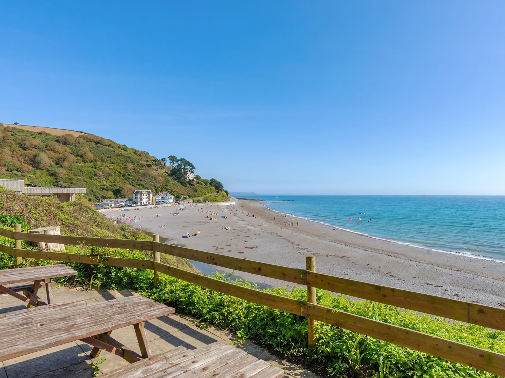 Ein Strand mit einer Holzpromenade, blauem Himmel und einer hügeligen Landschaft im Hintergrund.