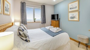 Ein Hotelzimmer mit einem Bett, einem Fernseher, einem Fenster mit Vorhängen, einer Pflanze und gerahmten Bildern an der Wand.