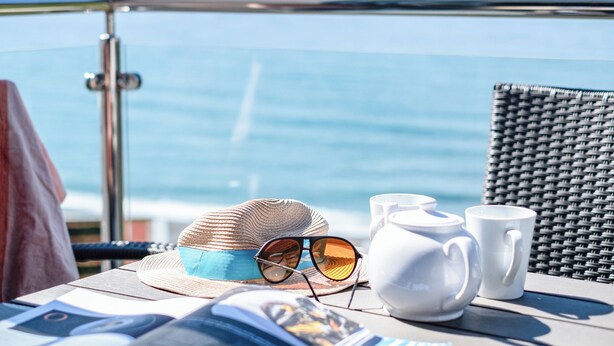 Ein Tisch mit einem Strohhut, Sonnenbrille und weißen Teetassen auf einem Balkon mit Blick aufs Meer.
