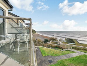 A balcony with metal chairs and a glass door overlooking a beach with a rocky shoreline and a clear sky. - 2 bedroom accommodation in Seaton (Seaton)