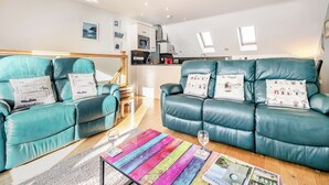 A modern living room with teal leather sofas, a colorful striped coffee table, and a kitchen in the background. - A contemporary house in a peaceful setting, with superb views over the sea. (Seaton)