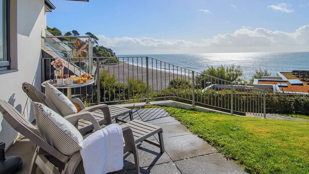 A balcony with lounge chairs, a small table with a vase of flowers, and a view of the beach and ocean. - A contemporary house in a peaceful setting, with superb views over the sea. (Seaton)