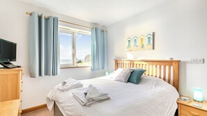 A neatly made bed with towels, a bedside table with a lamp, a TV on a wooden stand, and a window with curtains. - A contemporary house in a peaceful setting, with superb views over the sea. (Seaton)