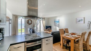 Een moderne keuken met een ingebouwde oven, een eethoek met rieten stoelen, en uitzicht op zee door grote ramen.