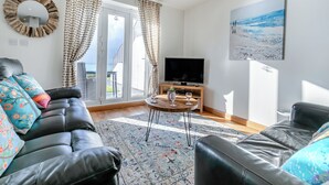 Een woonkamer met een zwarte leren bank, een houten salontafel en een televisie. Er is een groot raam met gordijnen, een schilderij aan de muur en uitzicht op het buitenland.