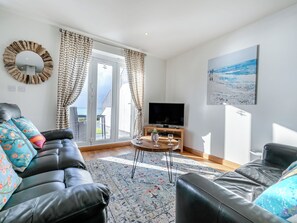 A living room with a black leather sofa, a wooden coffee table, and a television. There is a large window with curtains, a painting on the wall, and a view of the outdoors. - 24 Mount Brioni, Nr Looe (Seaton)