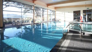 Una piscina interior con paredes de vidrio transparente, vistas al exterior y una estructura de techo de madera.