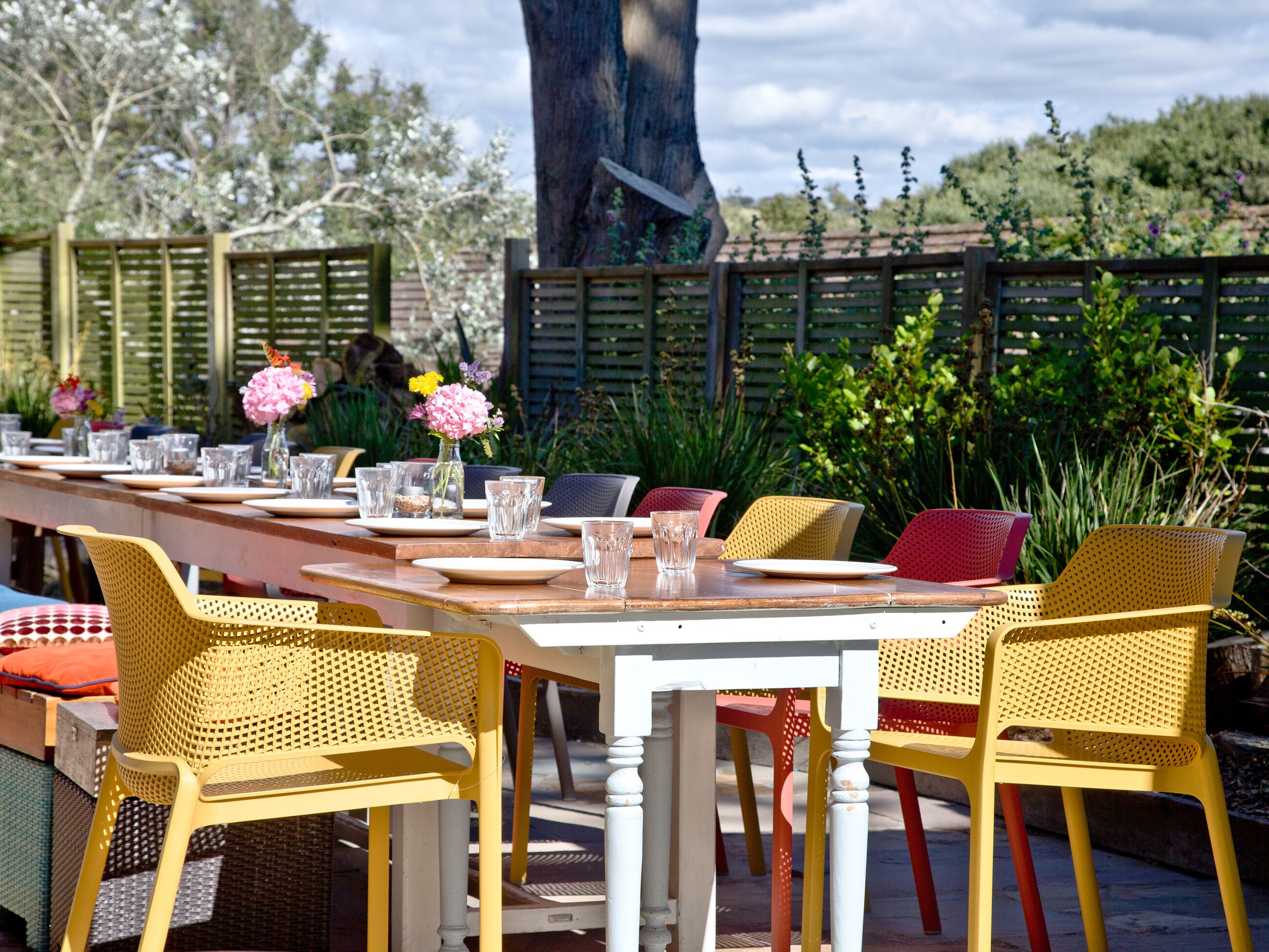 Un espace extérieur aménagé pour les repas, avec une longue table en bois dressée avec des assiettes, des verres et des fleurs. Des chaises jaunes et rouges sont disposées autour de la table.