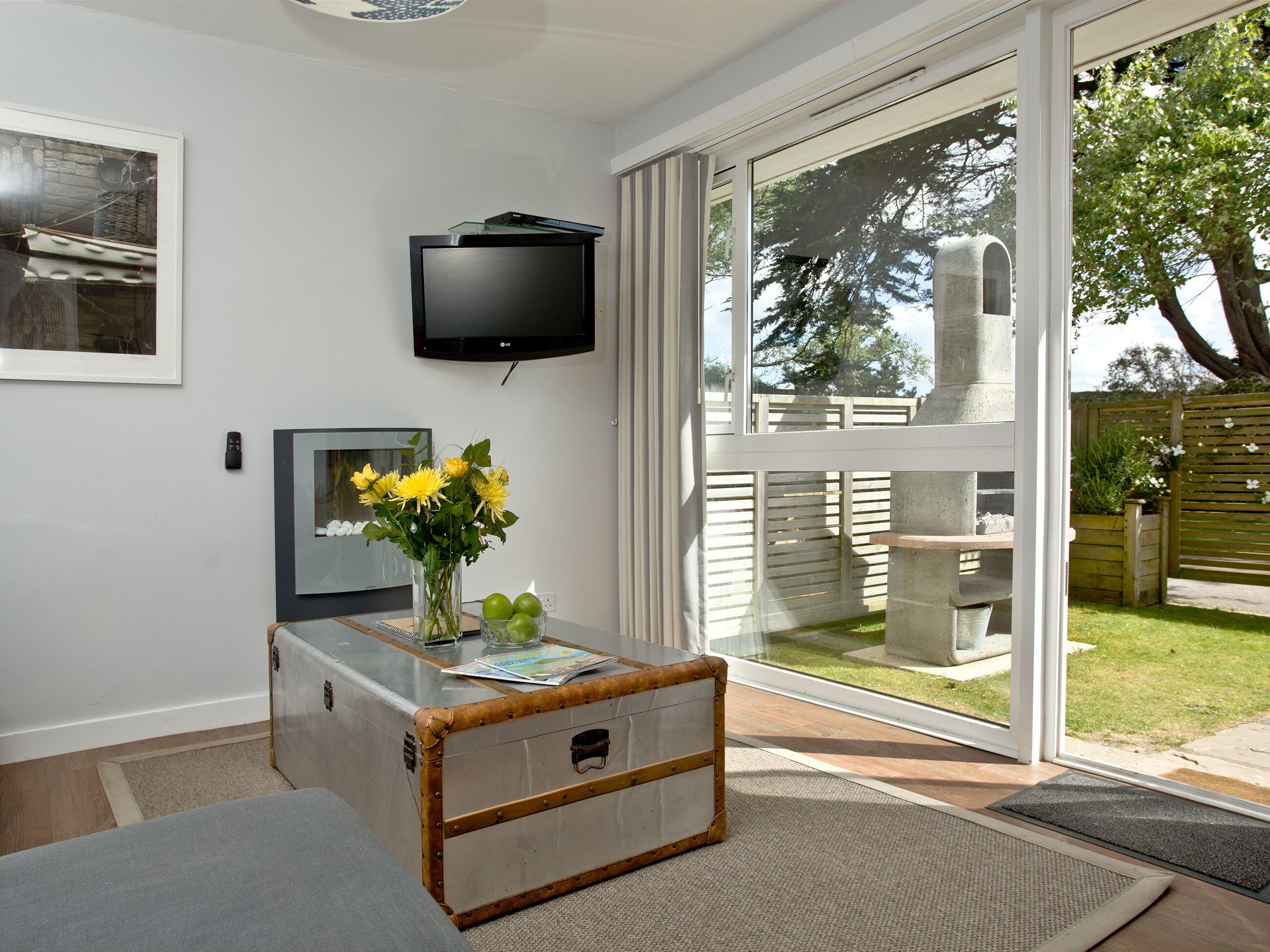 A modern living room with a glass door leading to a backyard, a flat-screen TV mounted on the wall, and a vintage-style table with a vase of flowers.