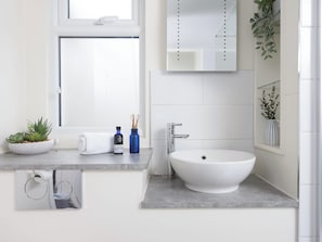 A bathroom with a white sink, a mirror, a window, a towel, a plant, and a bottle. - Little Dunley Cottages - Willow (Bovey Tracey)