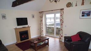 A living room with a fireplace, a television, and a large window with curtains. - Byre Cottages**Nr Stokesley (Carlton near Stokesley)