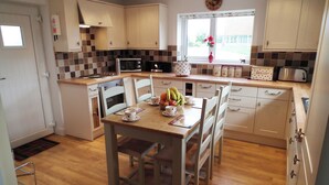 A kitchen with wooden cabinets, a dining table with chairs, a fruit bowl, and a window with a view of the outside. - Byre Cottages**Nr Stokesley (Carlton near Stokesley)