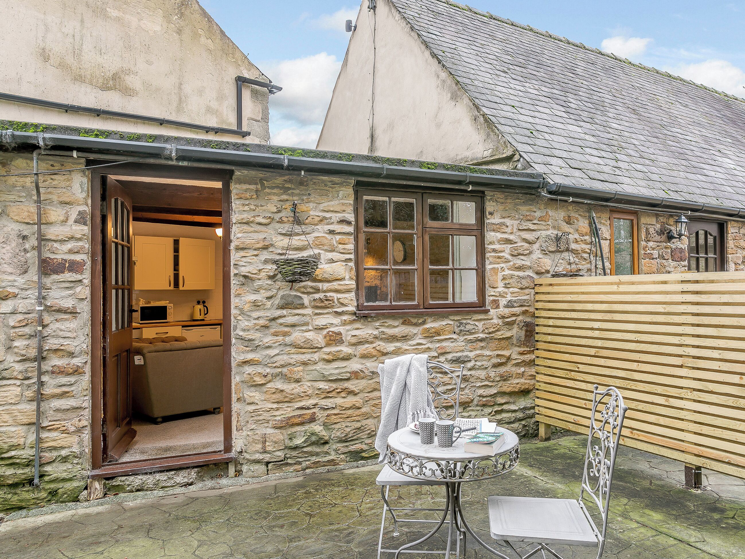 A stone building with a kitchen visible through an open door, a small round table with chairs, and a wooden fence.
