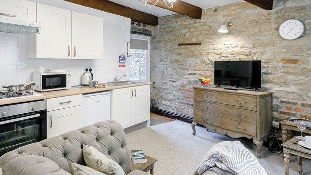 A modern kitchen with white cabinets, a microwave, and a stove. There's a stone wall with a clock and a wooden dresser with a TV.