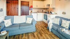 A cozy living room with two blue sofas, a wooden floor, and a kitchen area with white cabinets and appliances.