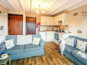 A cozy living room with two blue sofas, a wooden floor, and a kitchen area with white cabinets and appliances.