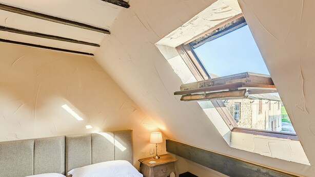 Una camera da letto con soffitto spiovente, una finestra a lucernario, un letto con lenzuola bianche, un comodino con una lampada e vista sugli edifici esterni.