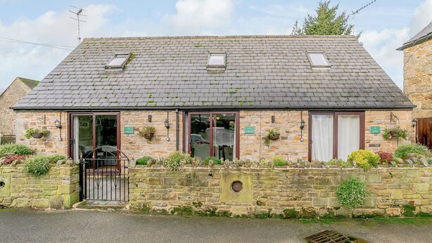 A stone building with a sloped roof, two windows with awnings, a gated entrance, and a small garden area with plants and flowers. - Mill Farm Cottages - Kingfisher (Barlow near Bakewell)