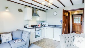 A compact kitchen with white cabinets, a built-in oven, and a microwave. There is a wooden ceiling with exposed beams and a chandelier. - Mill Farm Cottages - Kingfisher (Barlow near Bakewell)
