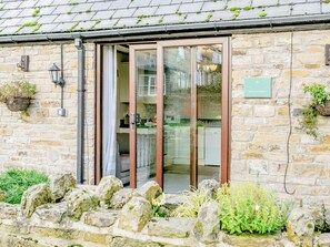 A stone building with a wooden door, a sign reading "KINGFISHER," and a stone wall with greenery. - Mill Farm Cottages - Kingfisher (Barlow near Bakewell)