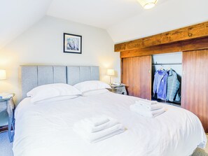 Een keurig opgemaakt bed met witte lakens en handdoeken, een houten hoofdbord, een ingelijst schilderij aan de muur en een houten garderobe met daarin hangende kleding.