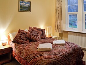 Een keurig opgemaakt bed met behang, een bedlampje, een stoel en een raam met gordijnen.