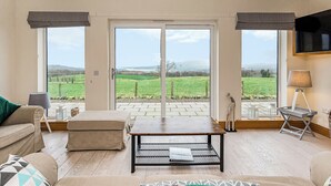 Una sala de estar con un sofá, una mesa de centro y un televisor. Amplias ventanas ofrecen vistas a un paisaje verde.