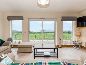 A living room with a sofa, a coffee table, and a TV. There are large windows with a view of a green landscape. - 2 bedroom accommodation in Gatehouse of Fleet (Gatehouse of Fleet)