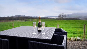 Una mesa con una botella de champán y dos copas, frente a un pintoresco paisaje de un campo verde y colinas lejanas.