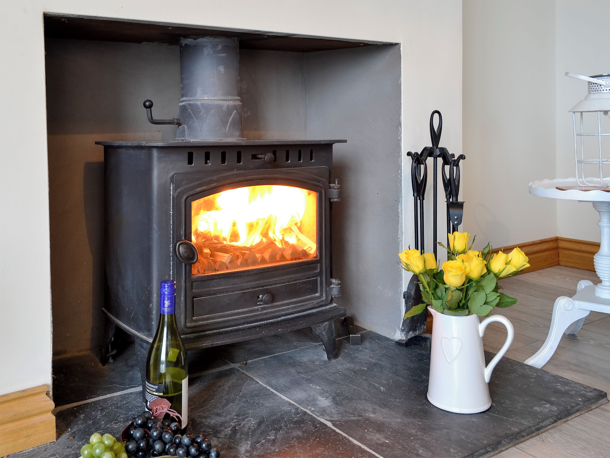 A wood-burning stove with a visible fire, a bottle of wine, a bowl of grapes, and a vase with yellow flowers.