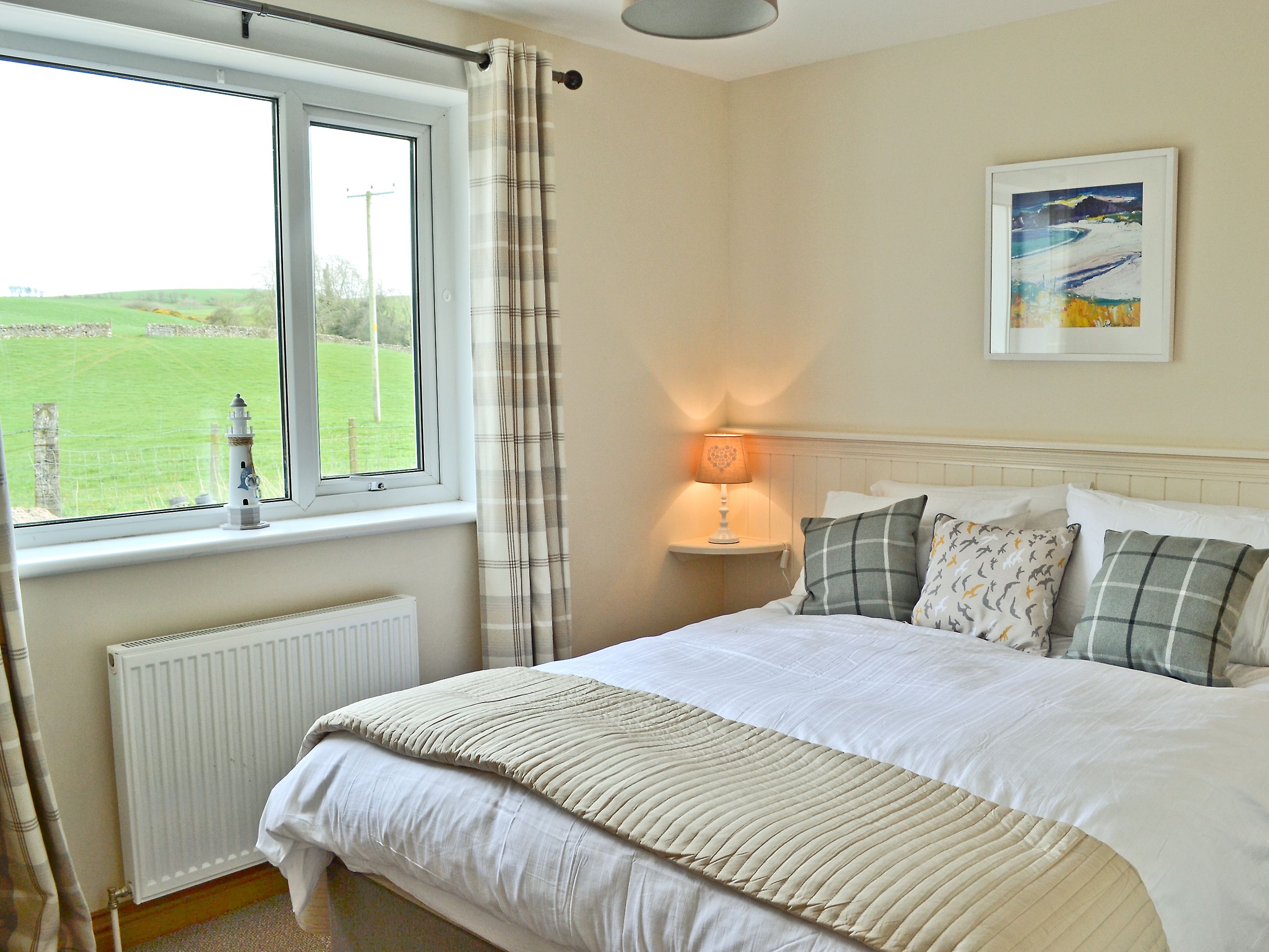 A bedroom with a large bed, a bedside table with a lamp, a window with curtains, a radiator, and a painting on the wall.