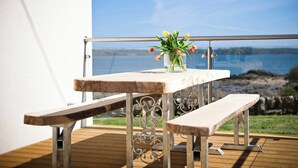 Ein Holztisch mit Blumen in einer Vase auf einem Balkon mit Metallgeländer, mit Blick auf ein Gewässer.