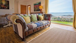 Ein gemütliches Wohnzimmer mit einem großen Fenster und Meerblick, einer bequemen Couch mit bunten Kissen und Holzboden.