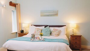 Una cama bien hecha con una colcha azul, dos mesitas de noche con lámparas, un cuadro en la pared y una ventana con cortinas.