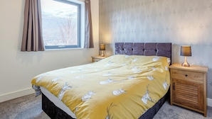 A bedroom with a bed, two bedside tables, a window with curtains, a patterned wall, and a nightstand with a lamp. - 5 bedroom accommodation in Kinlochleven (Kinlochleven)
