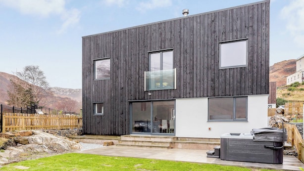 A modern two-story house with a dark wooden exterior, large windows, and a visible outdoor seating area. - 5 bedroom accommodation in Kinlochleven (Kinlochleven)