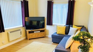 Ein Wohnzimmer mit einem Flachbildfernseher auf einem Holzständer, einem dunklen Sofa mit gelben Kissen, einer kleinen Topfpflanze und einem großen Teppich.