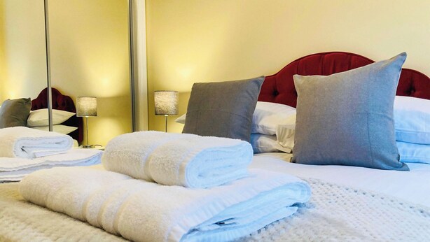 Ein ordentlich gemachtes Bett mit aufgerollten Handtüchern, einem roten Kopfteil und grauen Kissen.