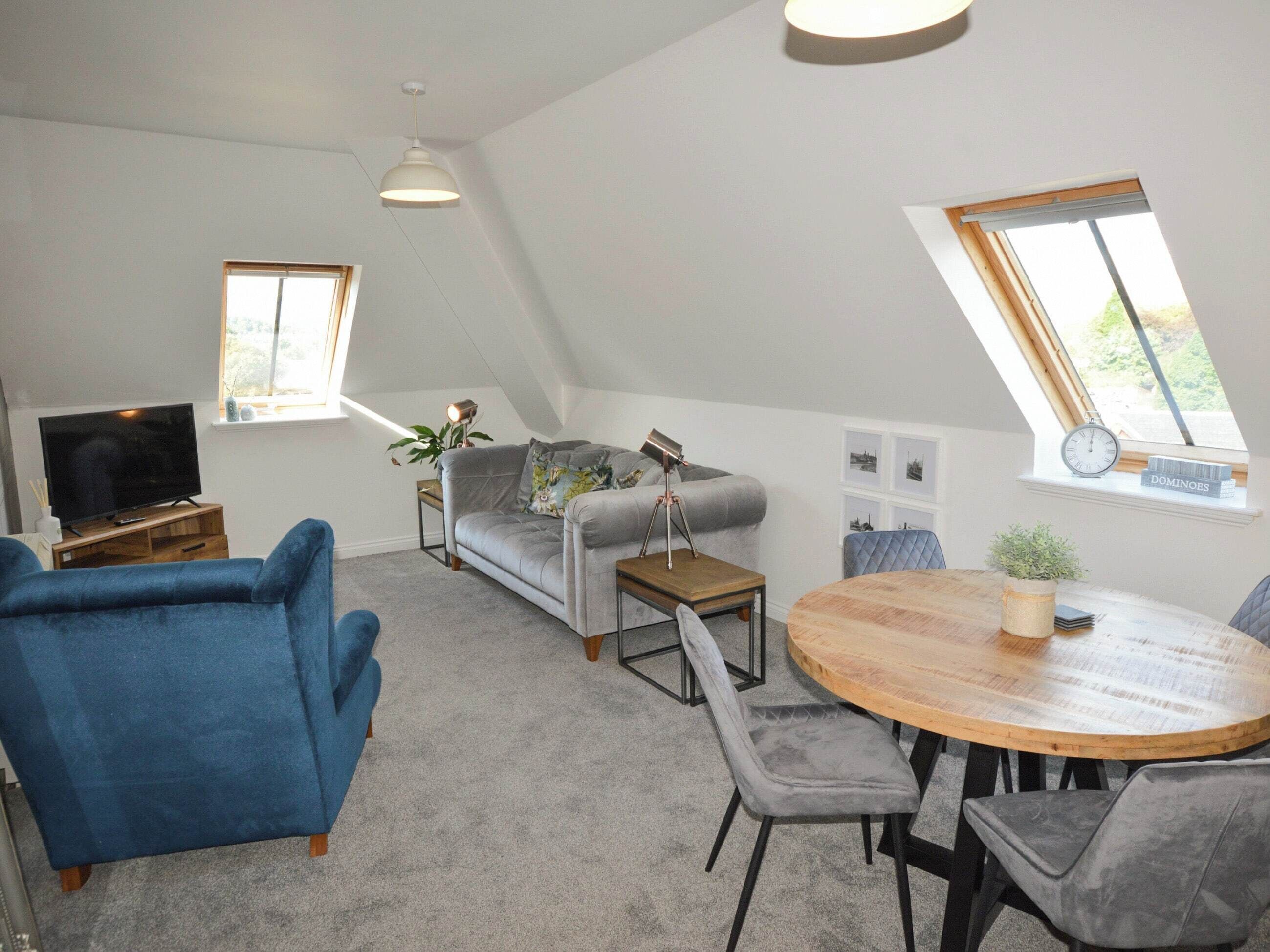 Uma sala de estar estilo loft com televisão de tela plana, sofá cinza, poltrona azul e uma mesa de jantar redonda de madeira.