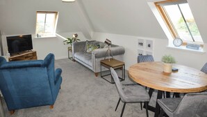 Ett vardagsrum i loftstil med en platt-TV, en grå soffa, en blå fåtölj och ett runt matbord i trä.