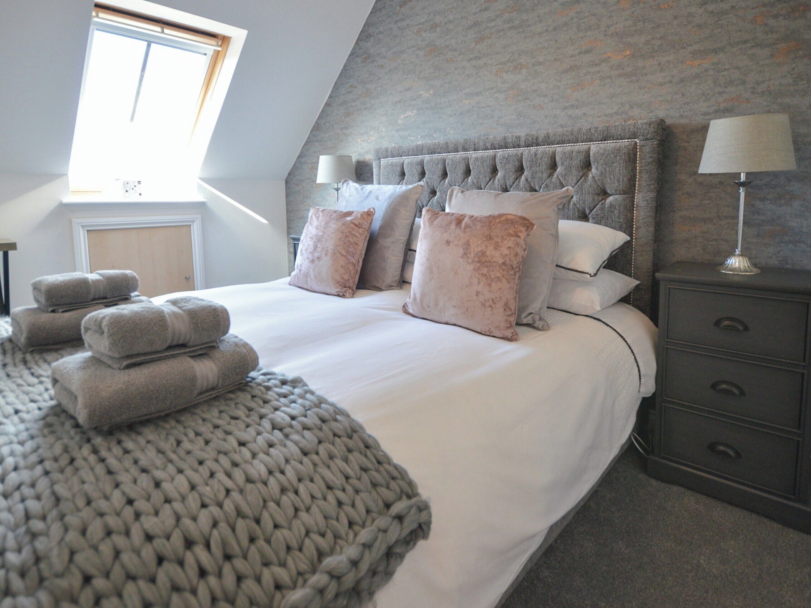 A bedroom with a large bed, multiple pillows, a grey knitted throw, a nightstand with a lamp, and a window with blinds.