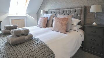 A bedroom with a large bed, multiple pillows, a grey knitted throw, a nightstand with a lamp, and a window with blinds.