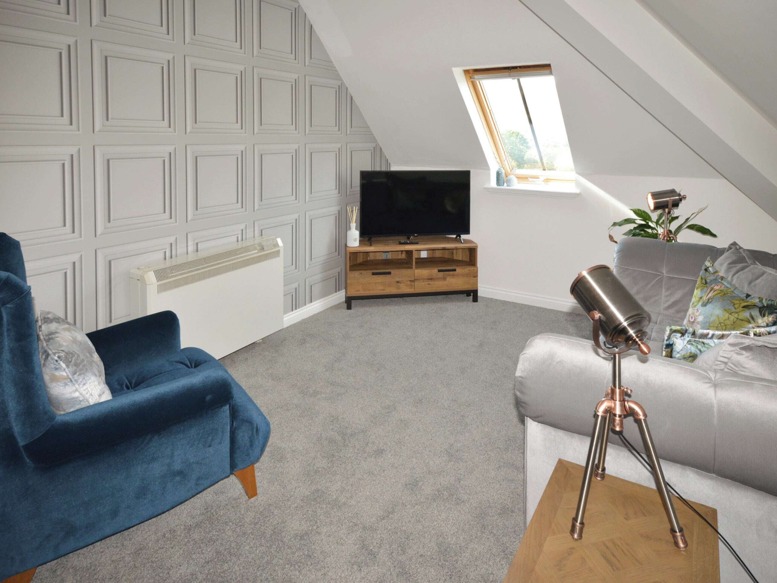 A modern loft bedroom with a sloped ceiling, a television on a wooden stand, a blue armchair, a grey sofa, and a telescope on a wooden table.