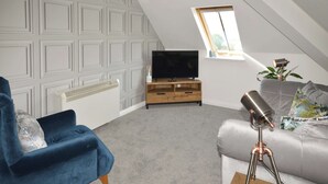 Ett modernt sovrum i loftstil med ett sluttande tak, en tv på ett träställ, en blå fåtölj, en grå soffa och ett teleskop på ett träbord.