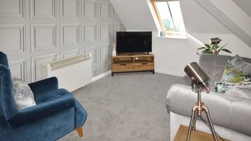 A modern loft bedroom with a sloped ceiling, a television on a wooden stand, a blue armchair, a grey sofa, and a telescope on a wooden table.