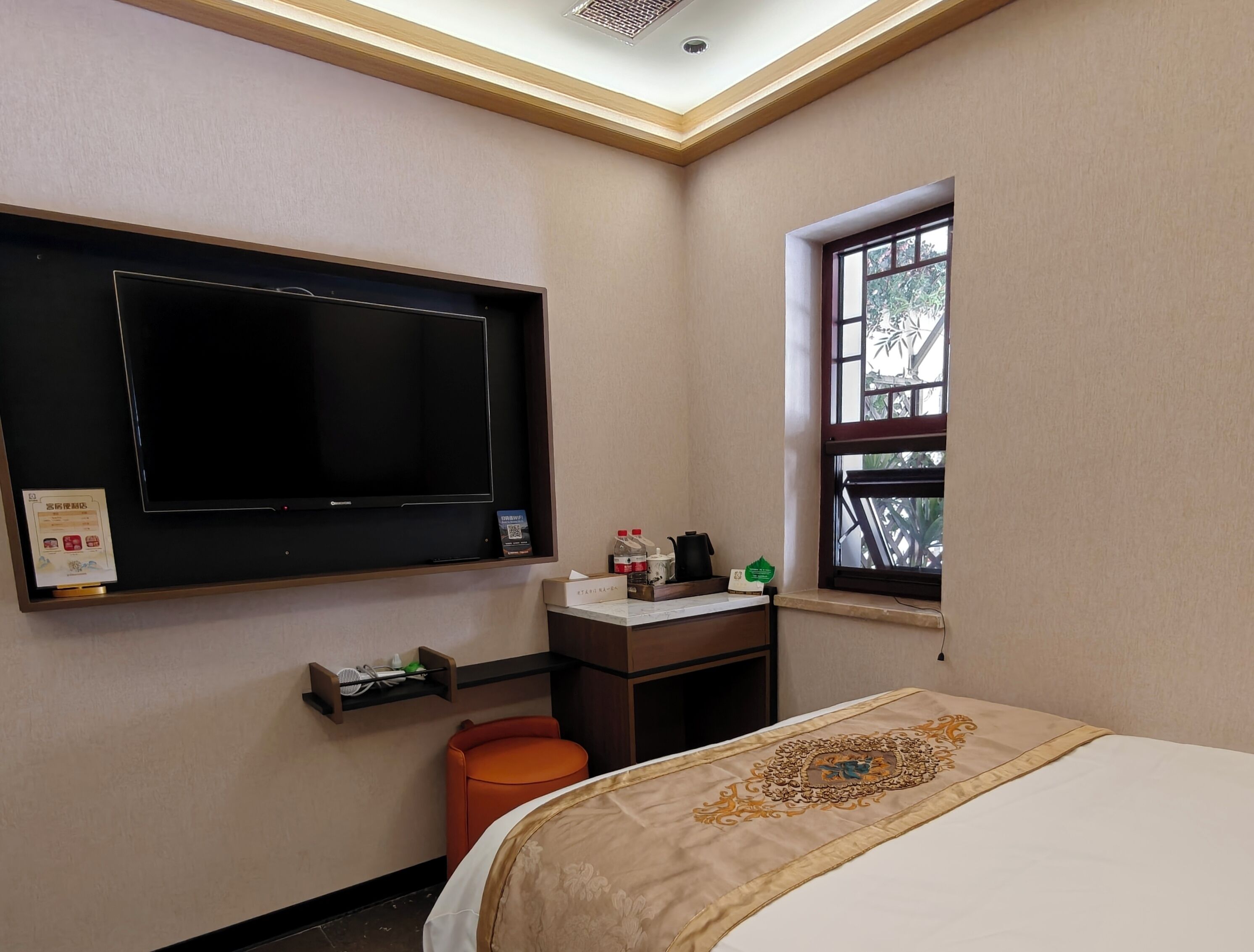 A hotel room with a flat-screen TV, a small desk with a chair, a window with a view, and a bed with a decorative headboard.