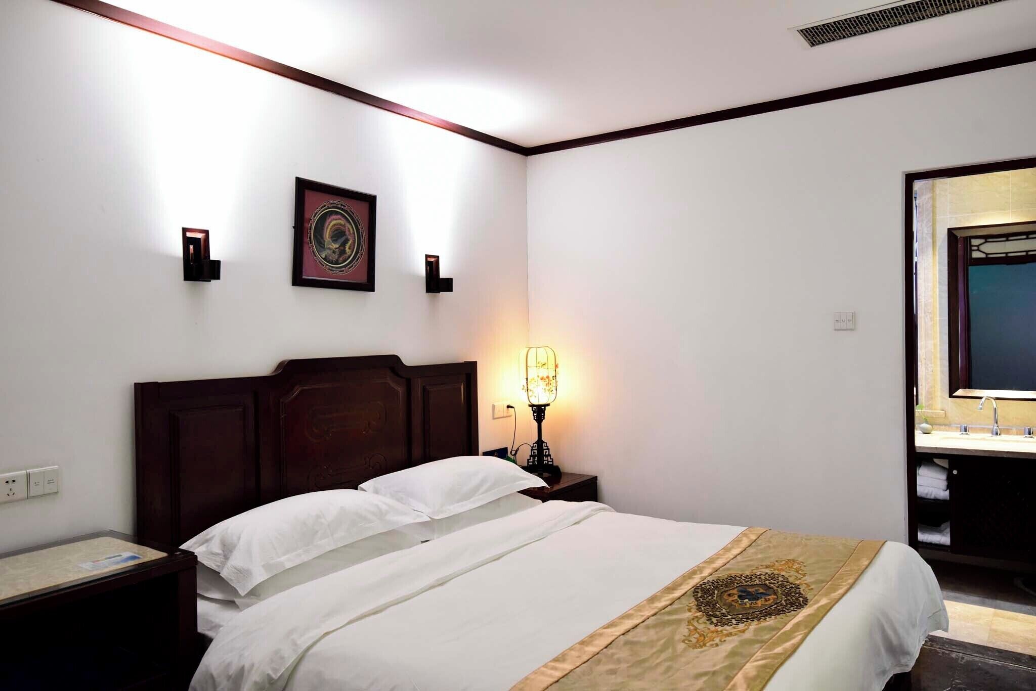 Une chambre d’hôtel avec une tête de lit en bois, une table de chevet avec une lampe, un tableau encadré au mur et une salle de bain visible à travers une porte ouverte.